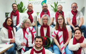Gruppe in Weiß mit roten Schals, hält Zuckerstangen und Adventskranz, festliche Stimmung.