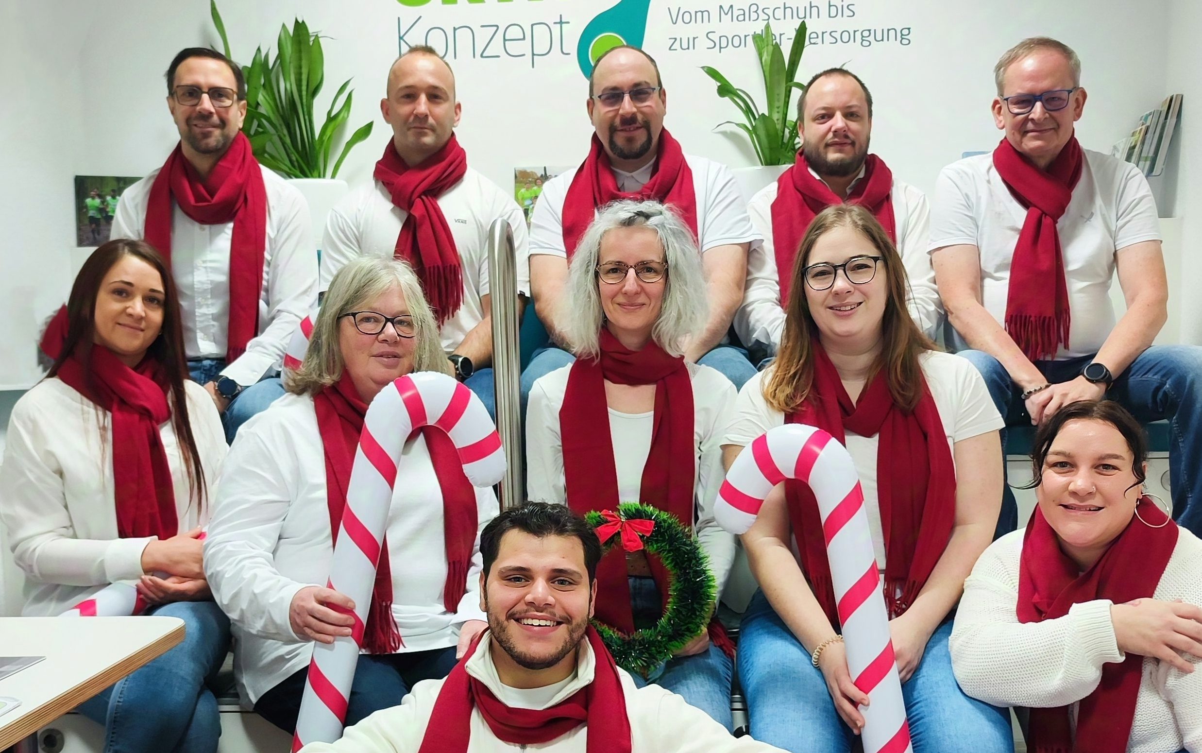 Gruppe in Weiß mit roten Schals, hält Zuckerstangen und Adventskranz, festliche Stimmung.